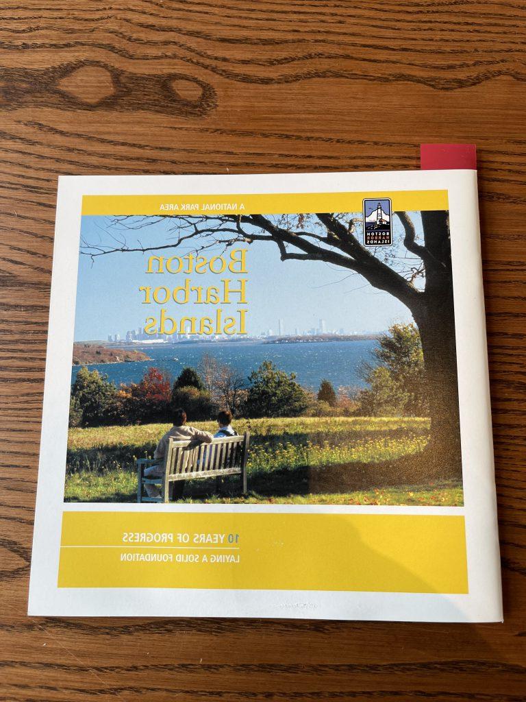 booklet with yellow borders and a picture of two people on a bench looking out onto the water with “Boston Harbor Island” written in yellow on the top right of the picture. In the bottom right corner of the booklet, it reads “10 years of progress/Laying a solid foundation” in white.]