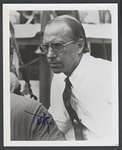 small black and white image of a white man with thinning hair wearing a white shirt with a dark tie. He is wearing large glasses on his face and is frowning.